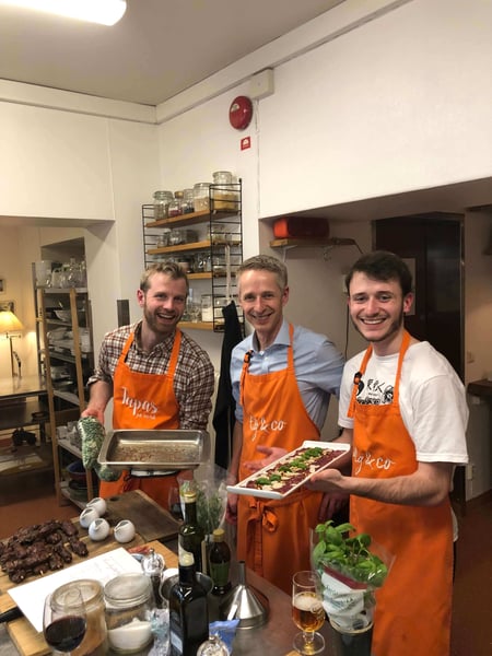 Three cheerful men in a kitchen, each wearing bright orange aprons, proudly presenting dishes they've prepared, with cooking ingredients and utensils around them.