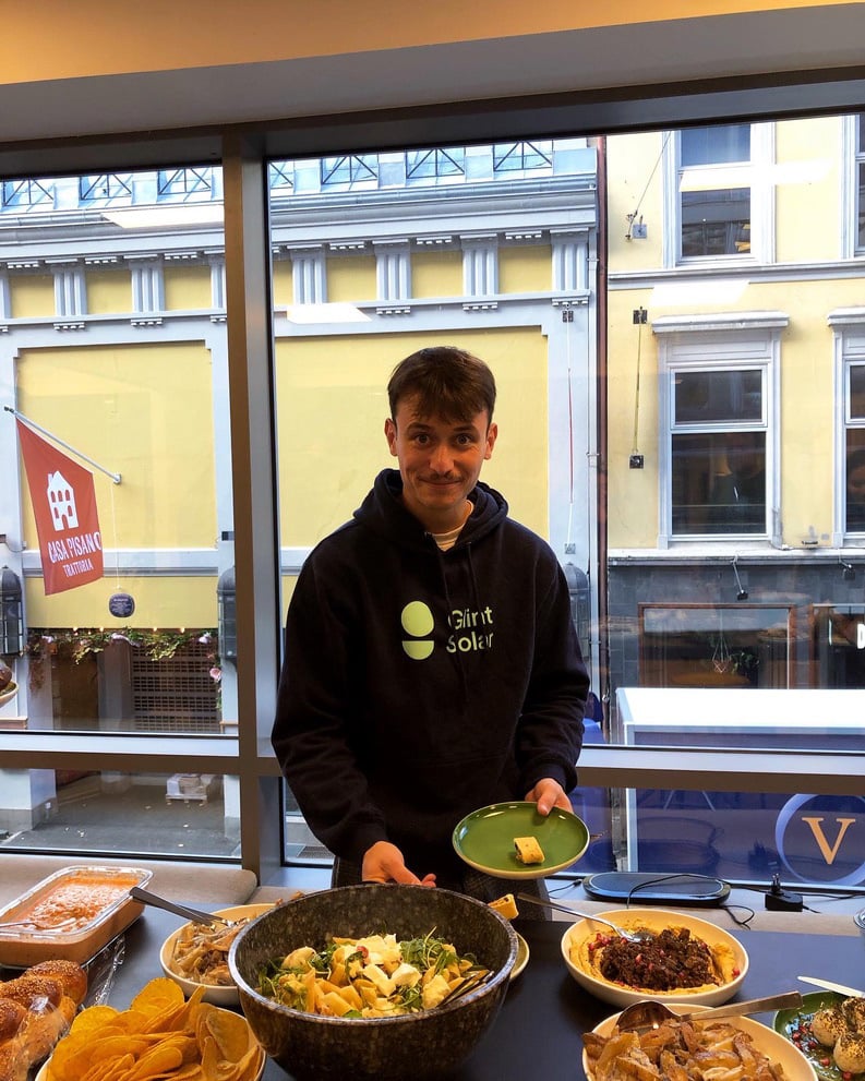 Martin, a team member, fills his plate with colorful food from a table while standing by a window in the office with a yellow building visible across the street during a team event.