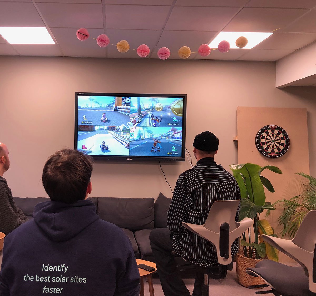 Three team members playing Mario Kart in front of a large TV. The photo, taken from behind, shows them in a relaxed setting with darts to the right and plants nearby.