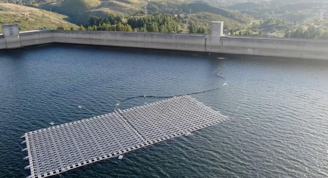 An aerial shot of a floating solar panel array on a tranquil reservoir, with a dam in the background amidst a hilly landscape.