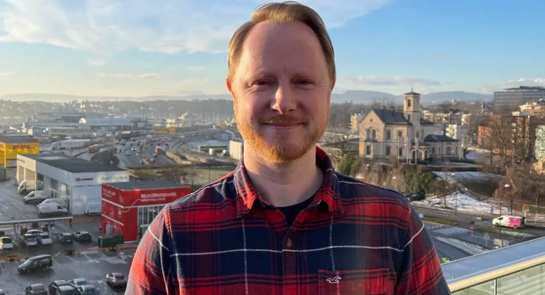 A man in a plaid shirt smiles at the camera, with a cityscape and clear blue skies in the background.